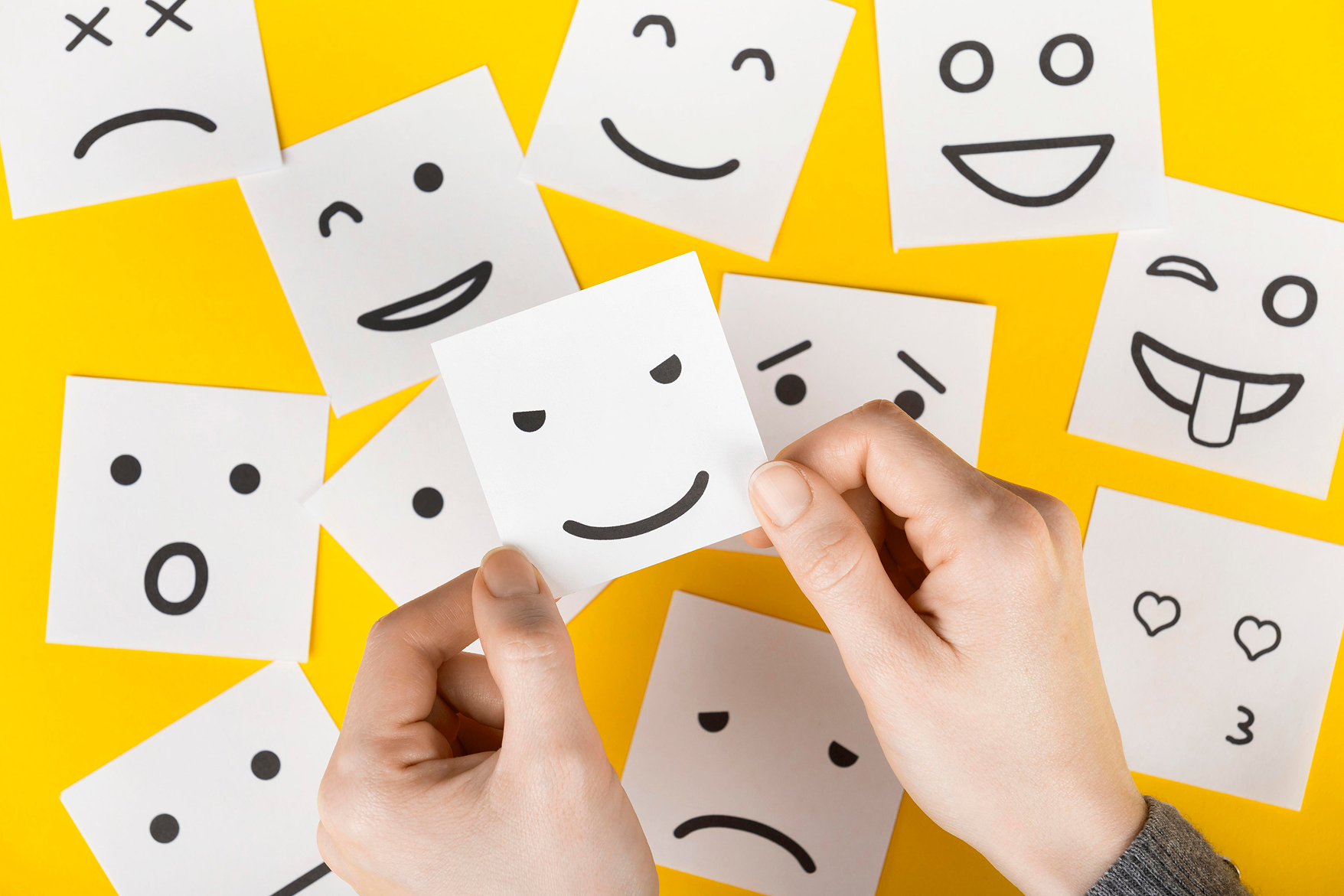Vue zenithal de plusieurs post-it avec des smileys de différentes émotions dessinées dessus. Ceux-ci sont posé sur un fond jaune.