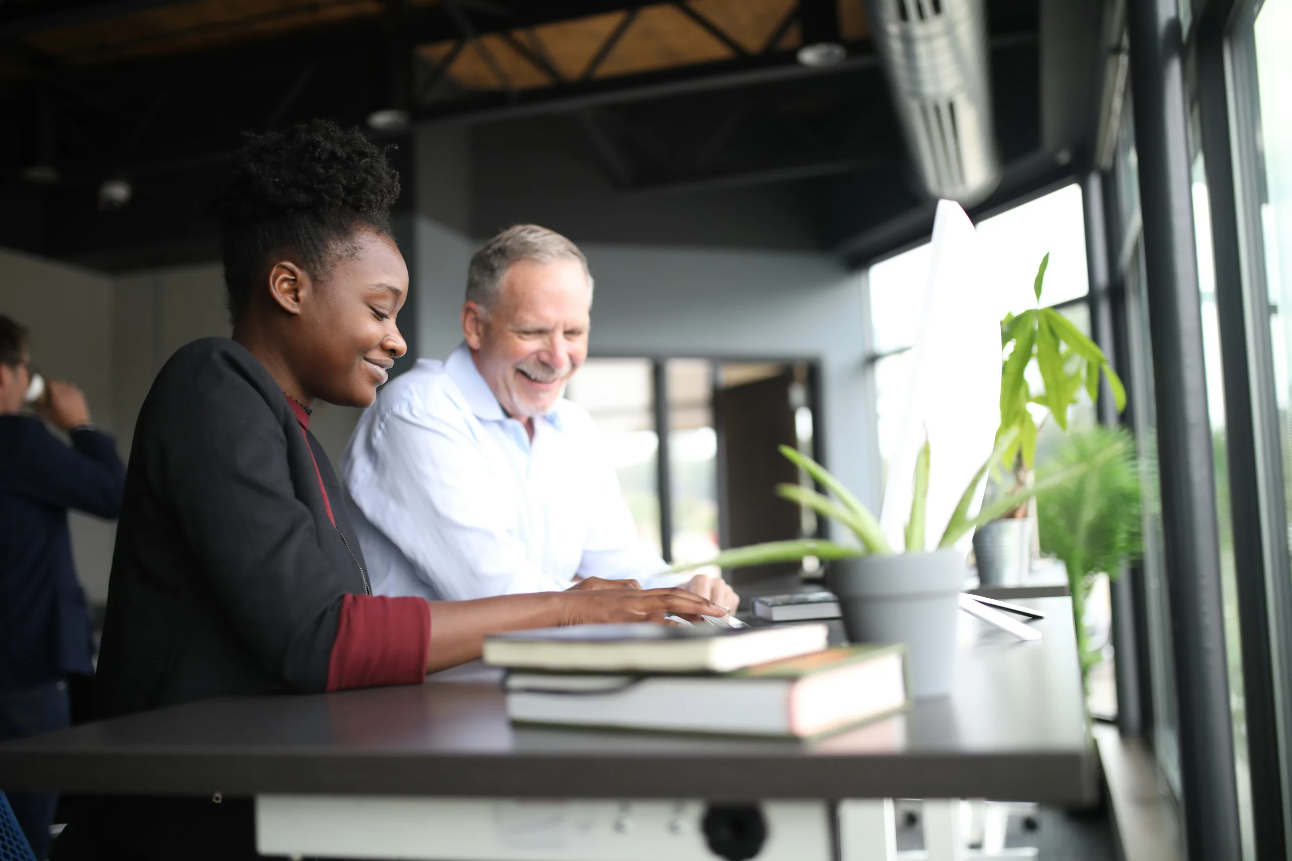 Une femme noire vêtu d'un blazer et d'une chemise bordeau (en premier plan) travaille un homme blanc plus âgé portant une chemise blanche. Ils arborent eux deux un sourire en regardant un document poser sur le bureau sur lequel ils se trouvent.